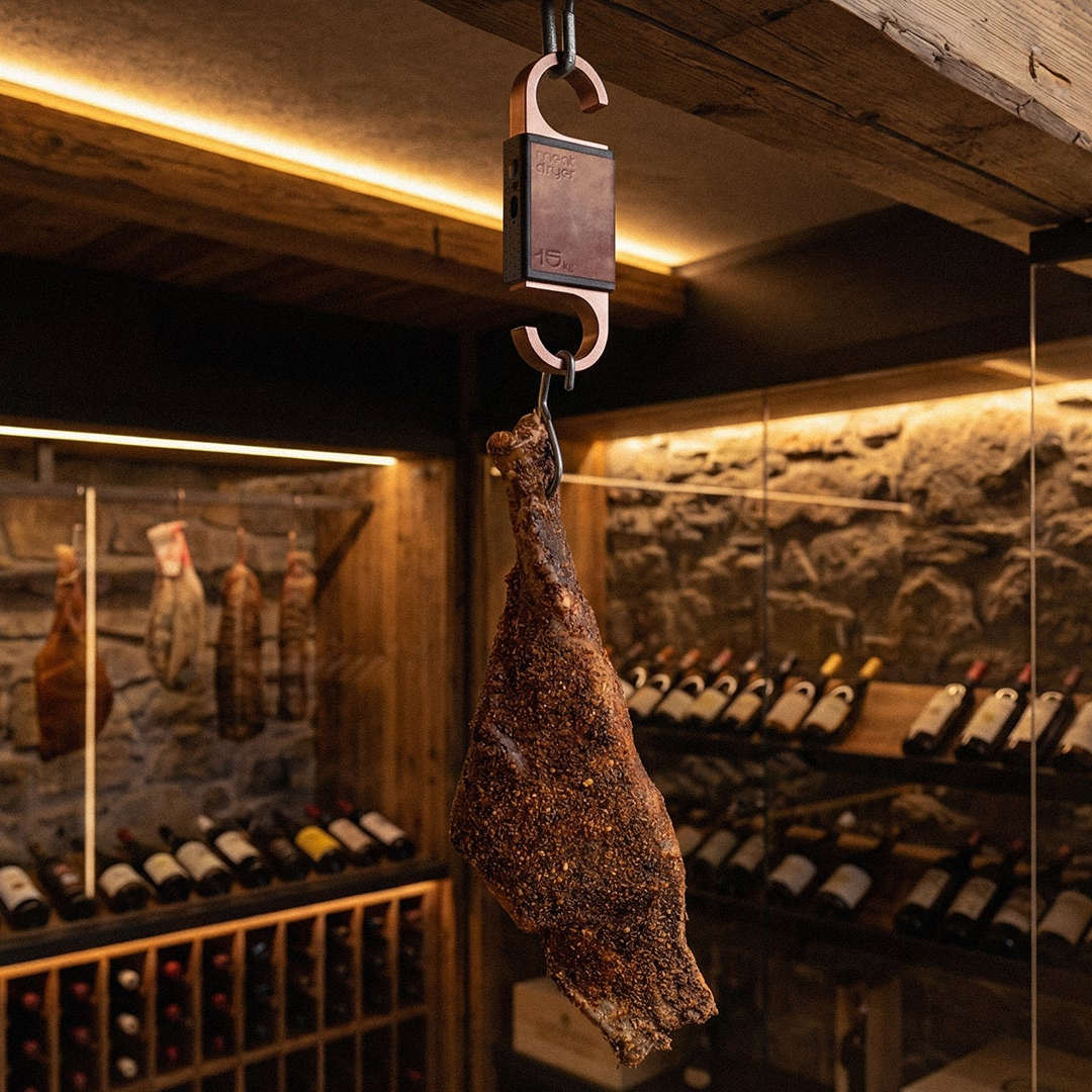 Cured meat hanging from a hook in an exclusive wine cellar with wine bottles and salami in the background.