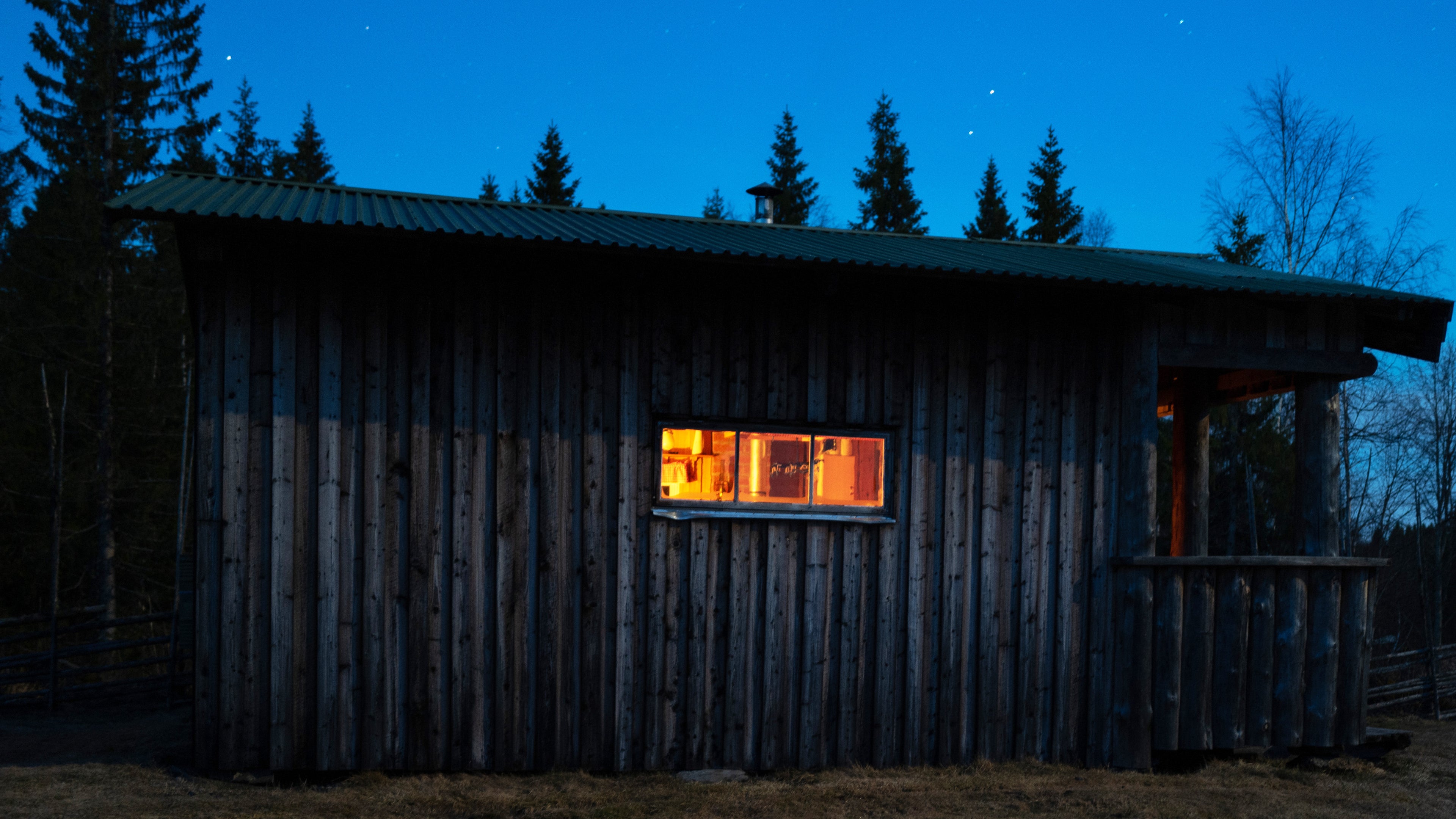 Wooden hunting cabin in a forest at night with one window illuminated.