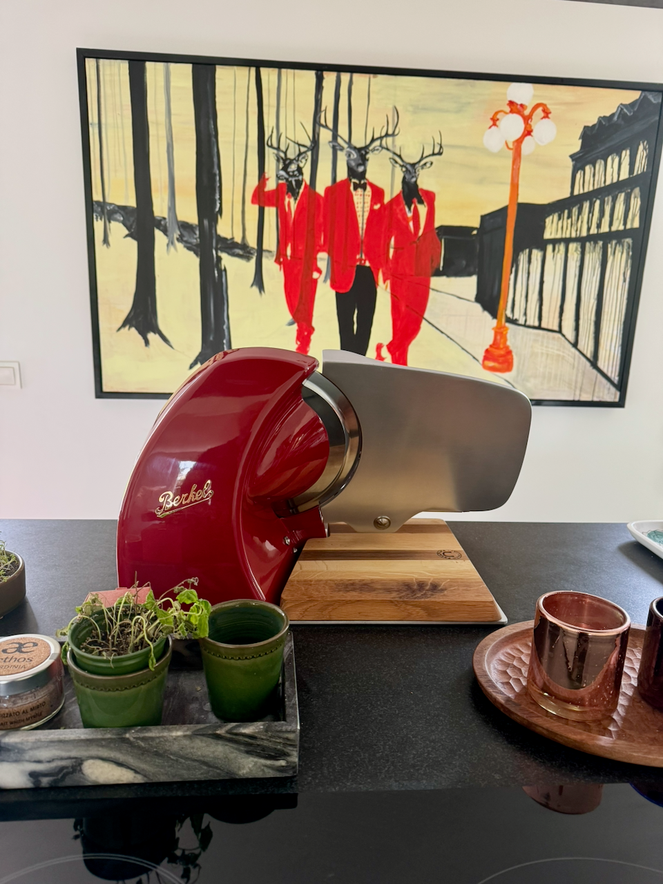 Red and silver Berkel meat slicer on a table with a painting of deer in red coats in the background.